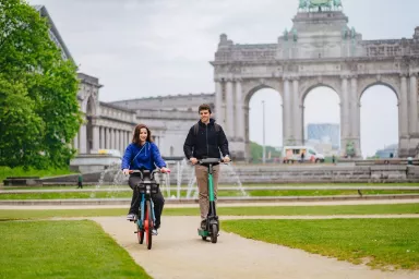 Une personne sur un vélo Dott et une personne sur une trottinette Bolt, devant les arcades du Cinquantenaire